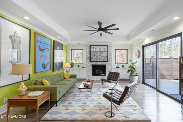 living room with light hardwood / wood-style flooring, a large fireplace, ornamental molding, ceiling fan, and a raised ceiling