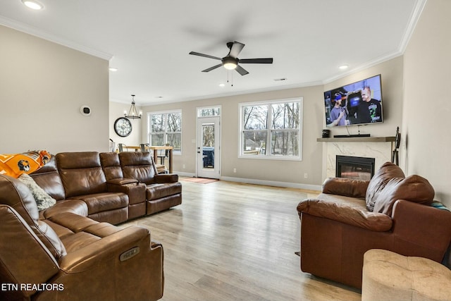 living room featuring crown molding, a high end fireplace, ceiling fan with notable chandelier, and light wood-type flooring