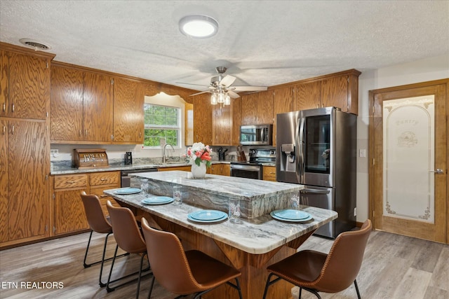 kitchen with light wood finished floors, appliances with stainless steel finishes, brown cabinetry, a sink, and a kitchen island