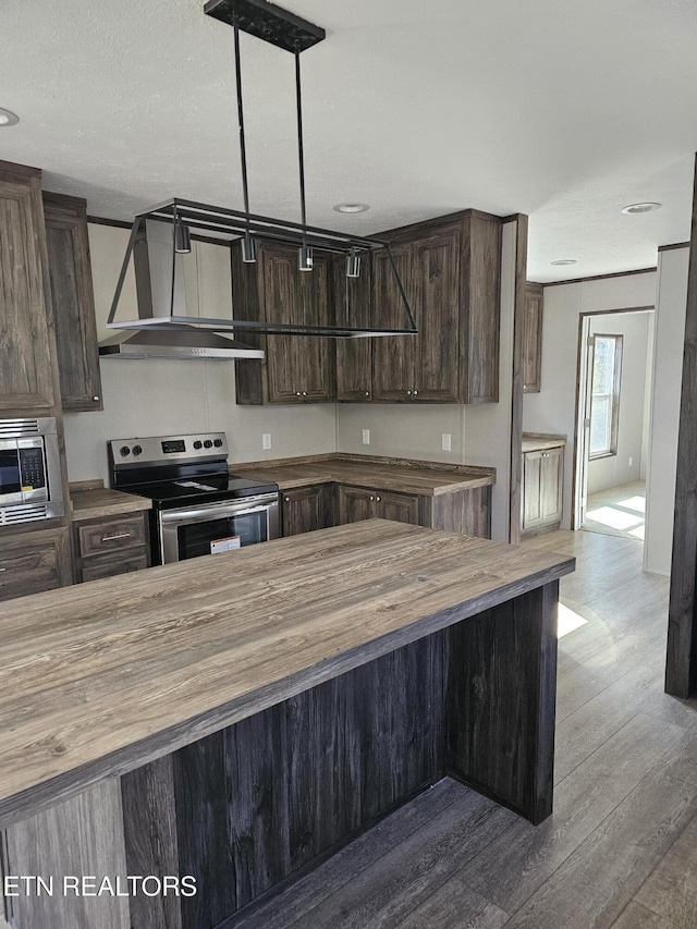 kitchen featuring appliances with stainless steel finishes, pendant lighting, wood counters, dark brown cabinets, and wall chimney exhaust hood