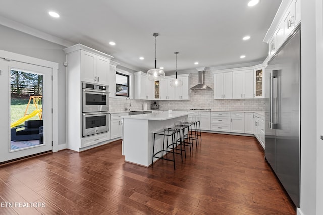 kitchen featuring a center island, appliances with stainless steel finishes, glass insert cabinets, a sink, and wall chimney range hood