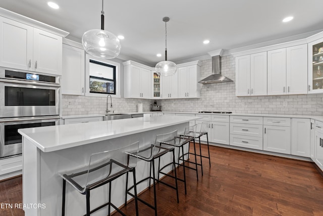 kitchen with wall chimney exhaust hood, stainless steel double oven, gas stovetop, white cabinetry, and a sink