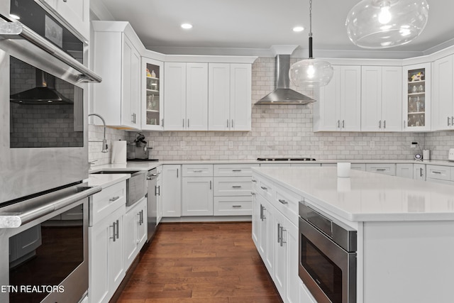 kitchen with dark wood-style floors, stainless steel appliances, light countertops, wall chimney range hood, and white cabinetry