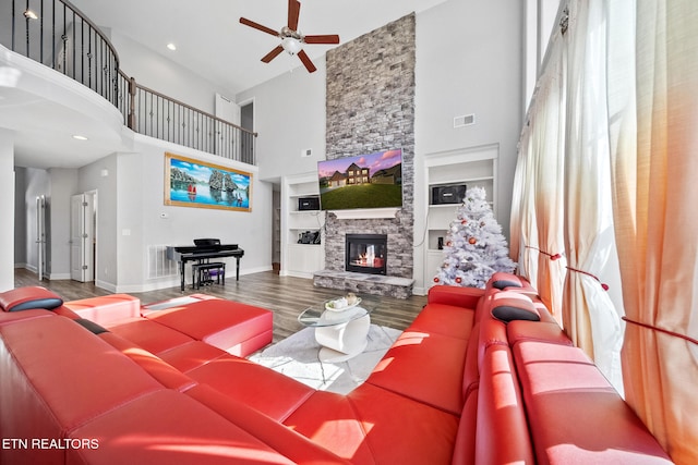 living room with a stone fireplace, wood-type flooring, a high ceiling, ceiling fan, and built in shelves