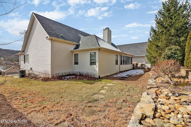 exterior space featuring central air condition unit, a shingled roof, a chimney, and a lawn