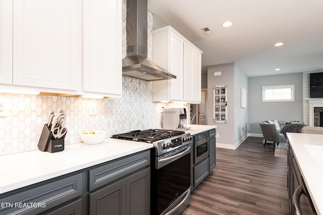 kitchen with appliances with stainless steel finishes, dark hardwood / wood-style floors, white cabinets, a brick fireplace, and wall chimney exhaust hood