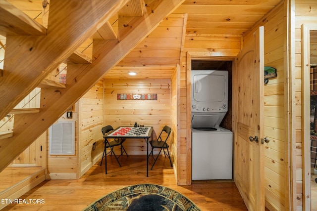 clothes washing area featuring hardwood / wood-style flooring, wooden ceiling, stacked washer and clothes dryer, and wood walls