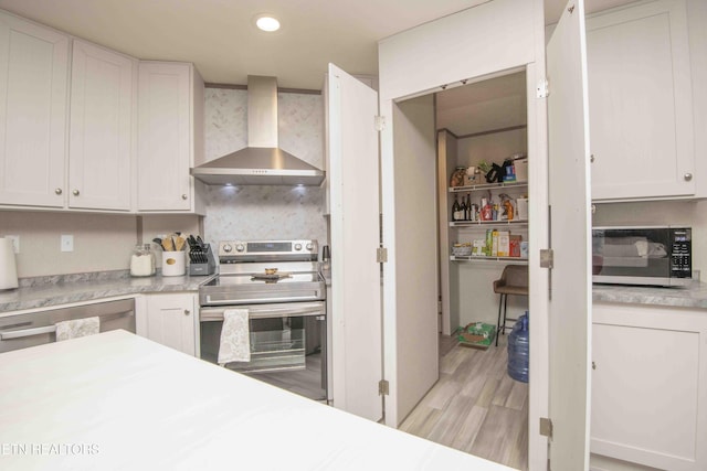 kitchen with wall chimney range hood, white cabinetry, light wood-style floors, appliances with stainless steel finishes, and light countertops