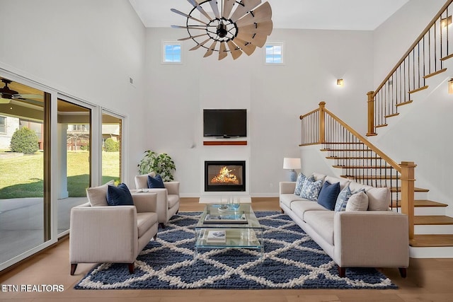 living area with a ceiling fan, a glass covered fireplace, wood finished floors, a towering ceiling, and stairs