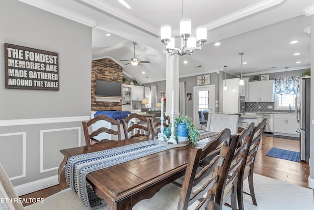 dining room with a stone fireplace, lofted ceiling, hardwood / wood-style floors, ceiling fan with notable chandelier, and ornamental molding