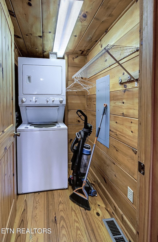 clothes washing area featuring electric panel, stacked washer / dryer, wood-type flooring, wooden ceiling, and wood walls