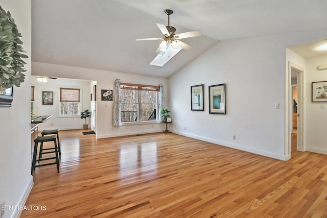 unfurnished living room with a ceiling fan, baseboards, lofted ceiling with skylight, and light wood finished floors