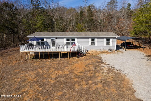 manufactured / mobile home with gravel driveway, a deck, a view of trees, and a detached carport