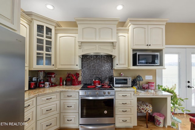 kitchen featuring cream cabinetry, premium range hood, backsplash, light stone counters, and appliances with stainless steel finishes