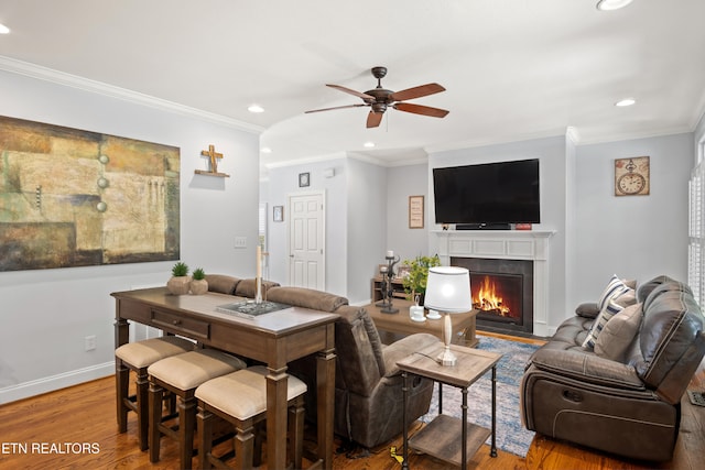 living area featuring ceiling fan, a fireplace with flush hearth, wood finished floors, baseboards, and crown molding