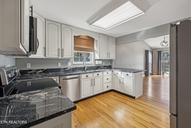 kitchen with a peninsula, a sink, appliances with stainless steel finishes, light wood-type flooring, and dark stone countertops