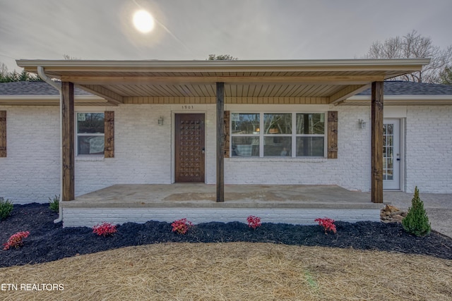 view of exterior entry with a porch, brick siding, and roof with shingles