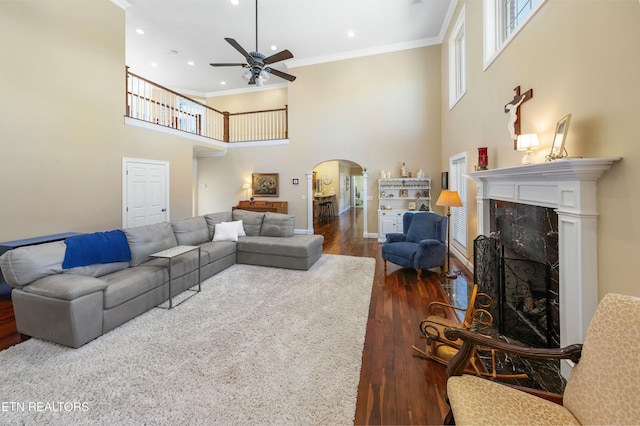 living area featuring arched walkways, dark wood-style flooring, crown molding, a fireplace, and a ceiling fan