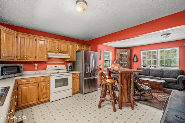 kitchen with under cabinet range hood, open floor plan, light countertops, appliances with stainless steel finishes, and brown cabinetry