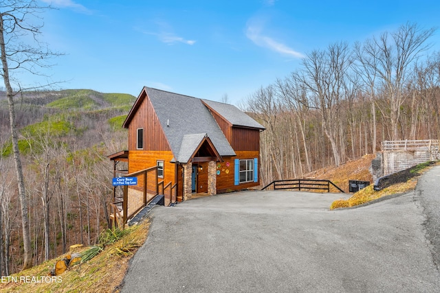 view of front of property featuring a shingled roof, driveway, a forest view, and fence