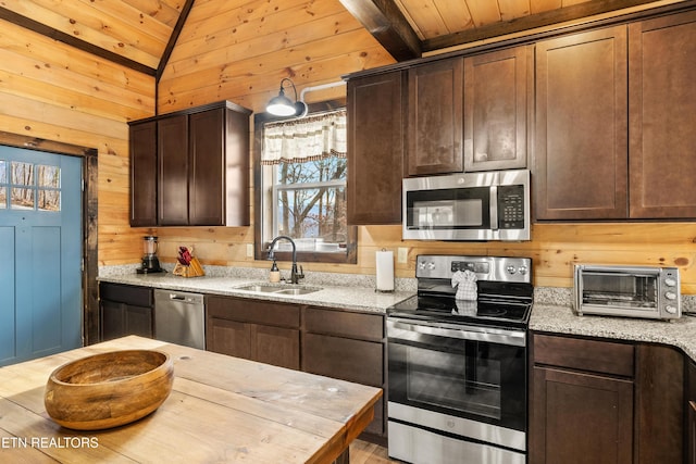 kitchen featuring lofted ceiling, light stone counters, wooden walls, stainless steel appliances, and a sink