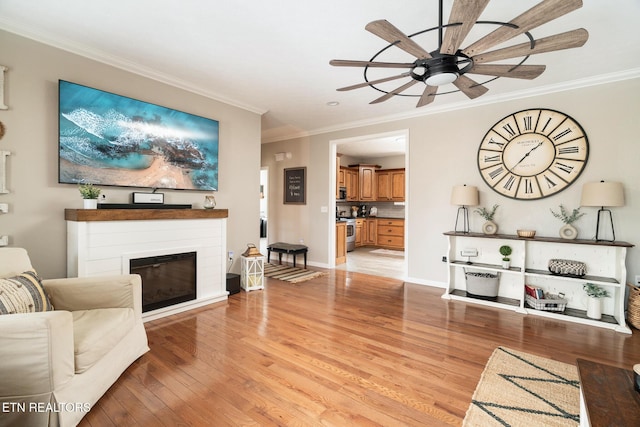 living area with light wood-style floors, a glass covered fireplace, ornamental molding, a ceiling fan, and baseboards