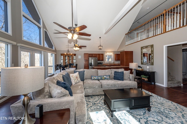 living room with high vaulted ceiling, ceiling fan with notable chandelier, baseboards, stairs, and dark wood finished floors