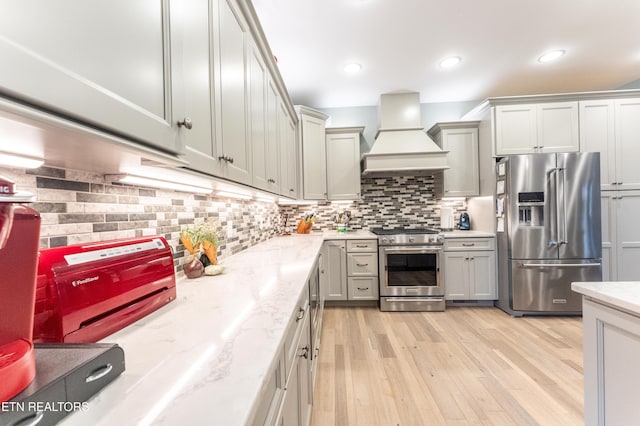 kitchen featuring premium appliances, custom exhaust hood, light wood-style flooring, decorative backsplash, and light stone countertops