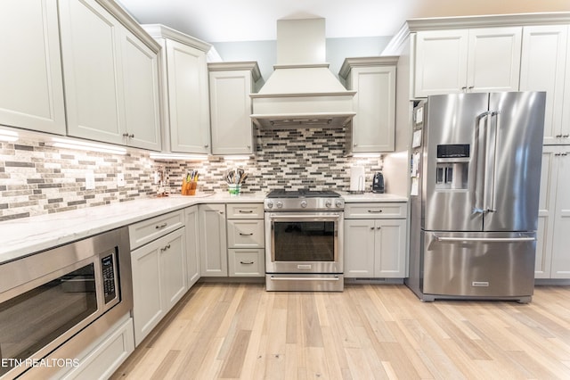 kitchen featuring light stone counters, light wood finished floors, tasteful backsplash, appliances with stainless steel finishes, and premium range hood