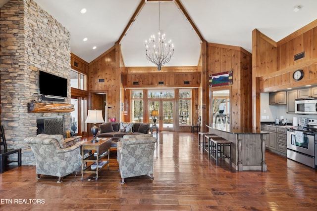 living room with dark wood finished floors, an inviting chandelier, a stone fireplace, wood walls, and high vaulted ceiling