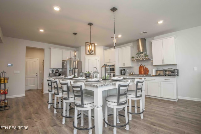 kitchen featuring an island with sink, wall chimney exhaust hood, appliances with stainless steel finishes, white cabinetry, and pendant lighting