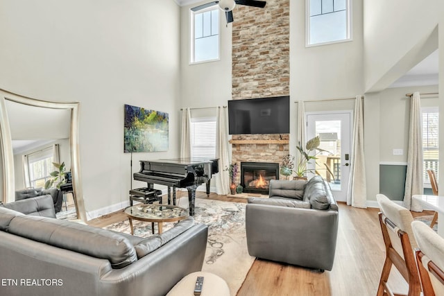 living room with a stone fireplace, light wood-style flooring, a high ceiling, a ceiling fan, and baseboards