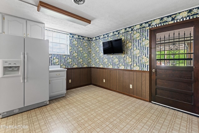 kitchen featuring a textured ceiling, light floors, white cabinetry, wainscoting, and white fridge with ice dispenser