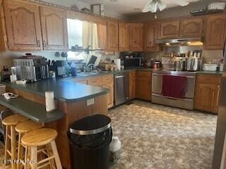 kitchen featuring a breakfast bar area, under cabinet range hood, a ceiling fan, appliances with stainless steel finishes, and brown cabinets