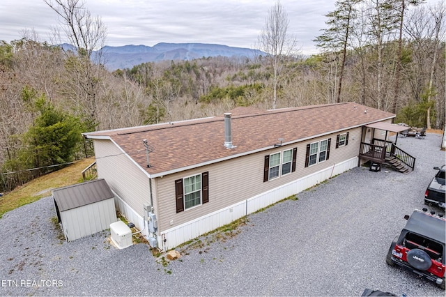 view of front facade featuring a mountain view, an outdoor structure, roof with shingles, gravel driveway, and a wooded view