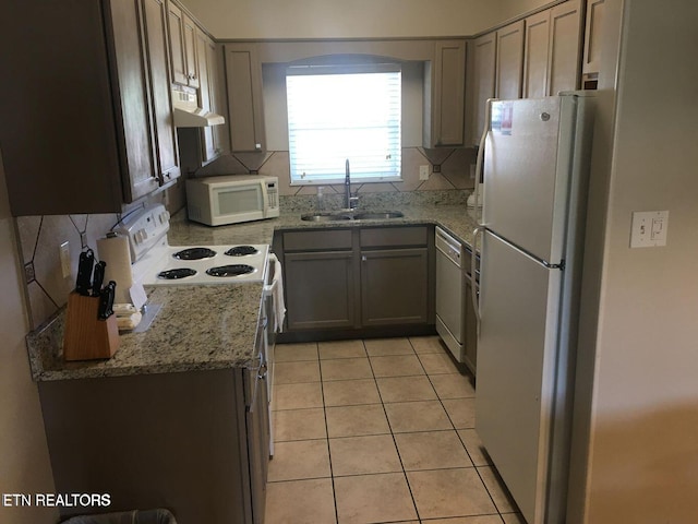 kitchen with light stone counters, light tile patterned flooring, under cabinet range hood, white appliances, and a sink