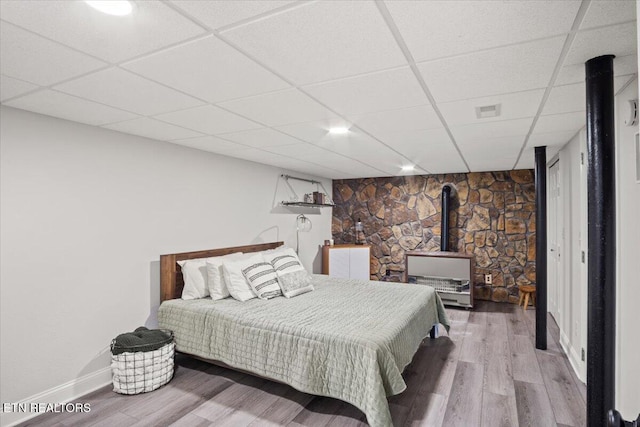 bedroom featuring a wood stove, baseboards, a drop ceiling, and wood finished floors