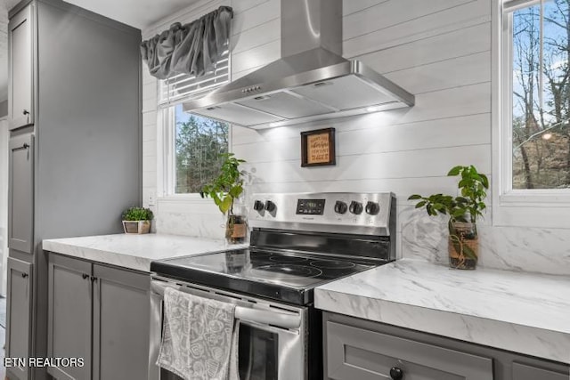 kitchen with wall chimney range hood, stainless steel electric range, light stone counters, and gray cabinetry