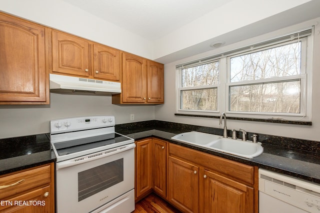 kitchen featuring brown cabinets, a sink, dark stone counters, white appliances, and under cabinet range hood