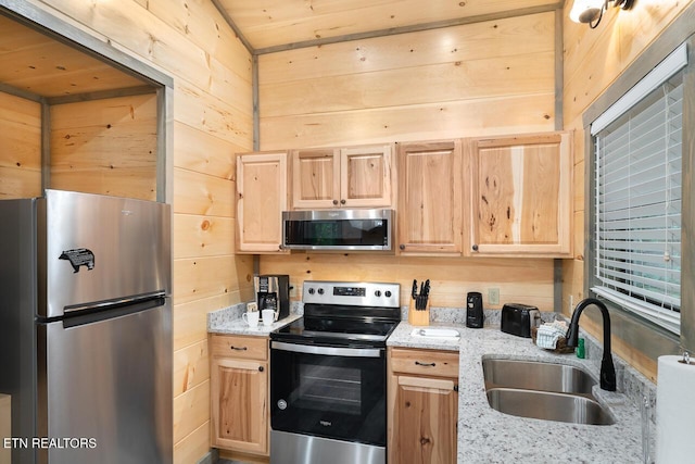 kitchen with appliances with stainless steel finishes, light brown cabinets, a sink, wood walls, and light stone countertops