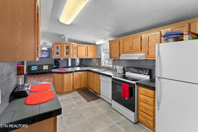 kitchen featuring under cabinet range hood, white appliances, a sink, dark countertops, and glass insert cabinets