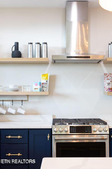 kitchen with light stone counters, open shelves, blue cabinets, stainless steel gas range oven, and wall chimney exhaust hood