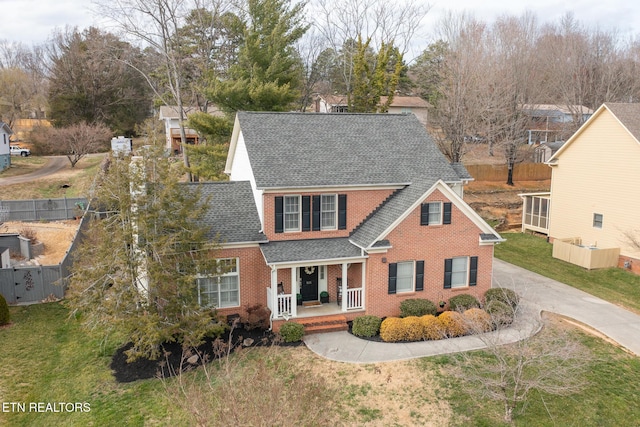 traditional-style house with a front yard, fence, roof with shingles, covered porch, and brick siding