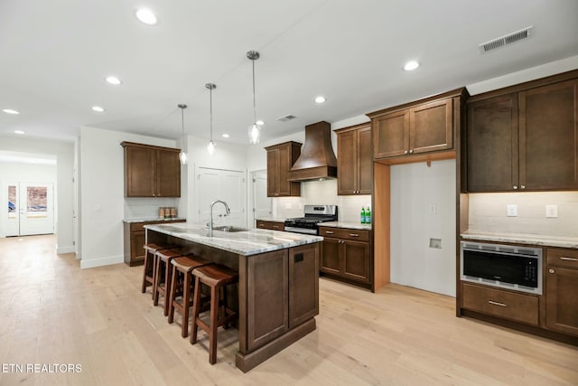 kitchen with light wood finished floors, visible vents, custom exhaust hood, stainless steel appliances, and a sink