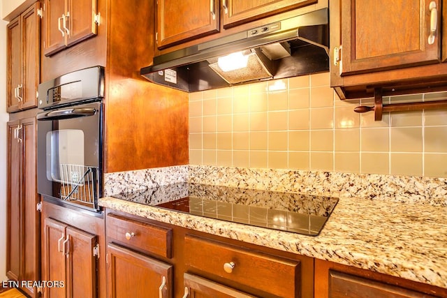 kitchen featuring decorative backsplash, light stone counters, brown cabinets, under cabinet range hood, and black appliances