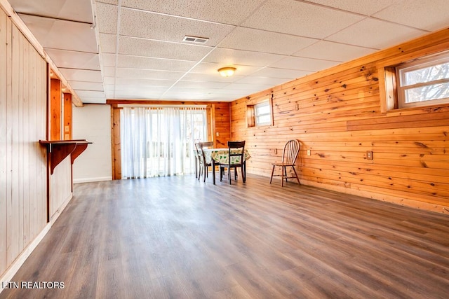 unfurnished dining area with wood walls, visible vents, a drop ceiling, and dark wood-style flooring