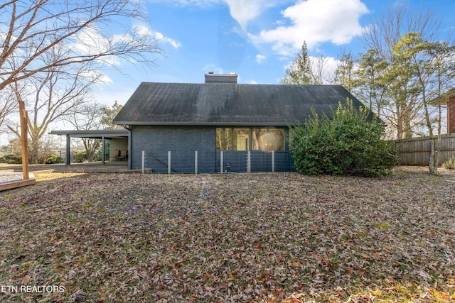 back of house featuring brick siding, a chimney, a shingled roof, fence, and an attached carport