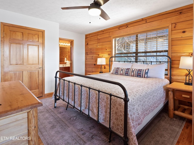 bedroom with a textured ceiling, connected bathroom, wooden walls, a ceiling fan, and baseboards