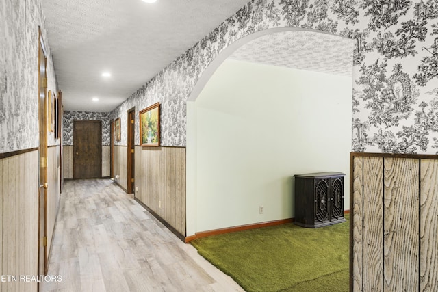 hallway with arched walkways, light wood-style floors, a textured ceiling, baseboards, and wallpapered walls