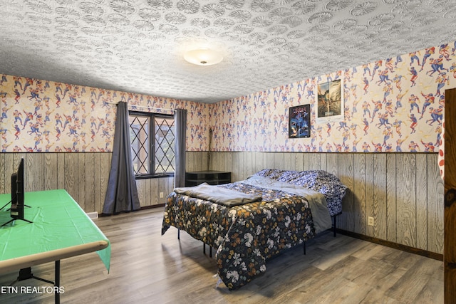 bedroom with a wainscoted wall, a textured ceiling, wood finished floors, and wallpapered walls